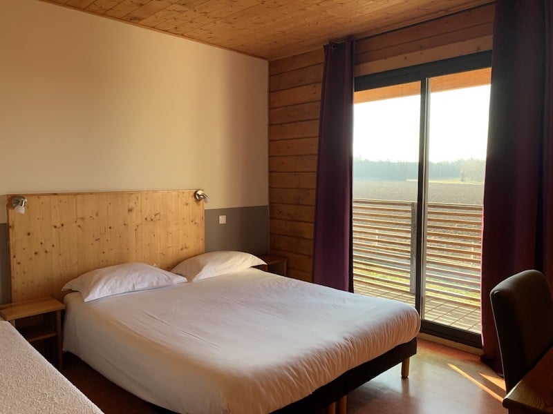 Chambre d'hôtel lumineuse avec un grand lit, tête de lit en bois clair, et une baie vitrée donnant sur un paysage de champs et un balcon à lattes horizontales.