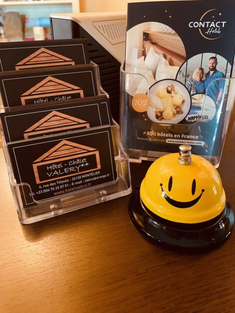 Comptoir d'hôtel avec porte-cartes Hôtel-Chalet Valéry, un dépliant Contact Hotels et une sonnette jaune souriante.