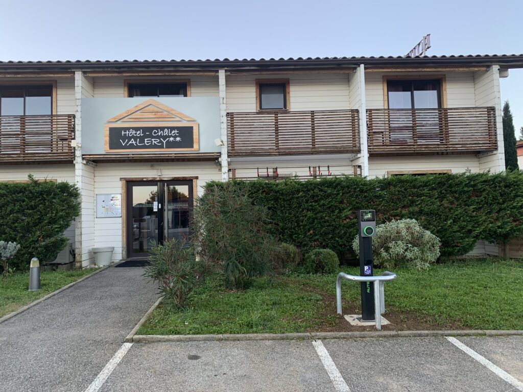 Façade de l'Hôtel - Chalet Valery** blanc avec balcons en bois, signalétique de borne de recharge EV et allée.