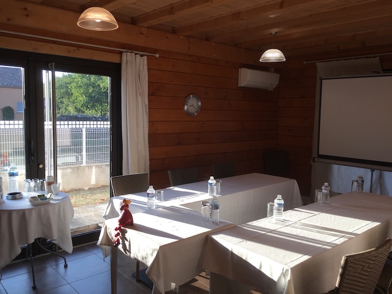 Salle de réunion ou de formation en bois, éclairée par la lumière du jour entrant par de larges portes vitrées, avec tables nappées et bouteilles d'eau.