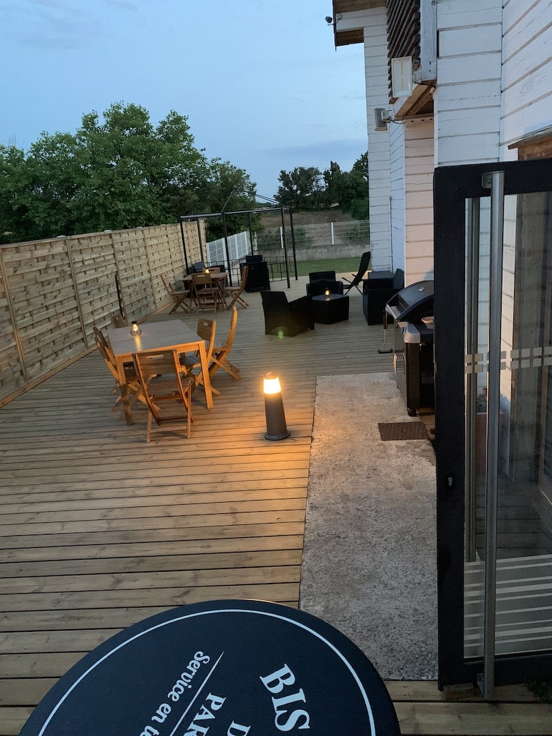 Terrasse en bois avec tables et chaises de jardin en soirée, éclairée par des lampes. Un panneau rond noir visible en bas porte l'inscription "BIS PAR... Service en te".